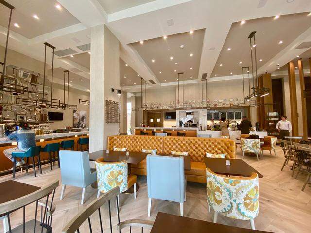The expansive dining room of a modern, brightly lit restaurant. In the center, a long, tufted banquette in a caramel leather provides seating for multiple tables. The room has high ceilings, a long bar to the left, and a full-view open kitchen along the back wall where chefs are working. The floors are a light wood laid in a herringbone pattern.